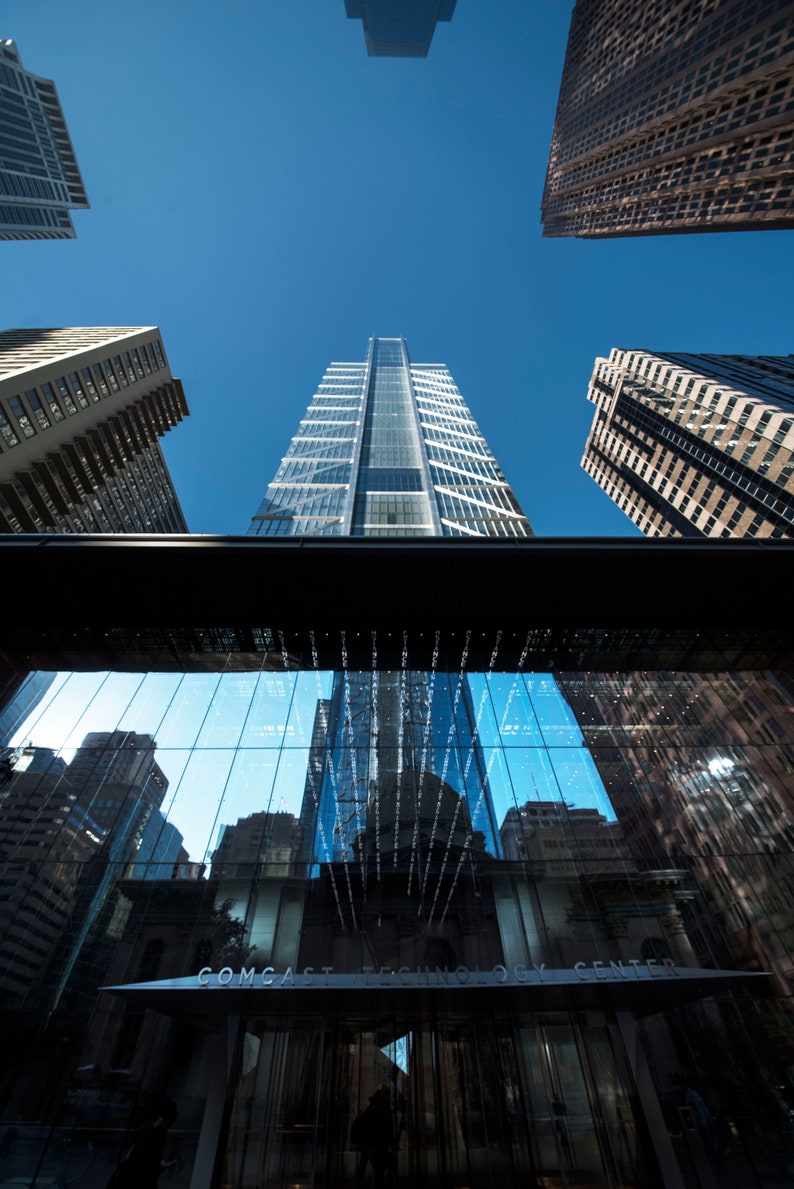 May include: A modern glass building with a reflective facade. The building is surrounded by other skyscrapers and the sky is blue. The building has a sign that reads "Comcast Technology Center".
