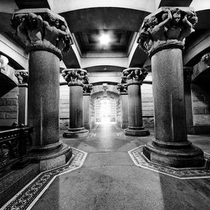 May include: A black and white photo of a hallway with a series of columns, each topped with a sculpture of multiple figures. The hallway is lit by a single light source at the end, creating a dramatic effect.