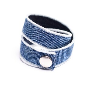 Può includere: Un braccialetto in denim blu con bordino bianco e chiusura a bottone argentata.