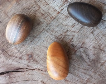 May include: Three wooden eggs, one dark brown, one light brown, and one medium brown, are arranged on a wooden surface.