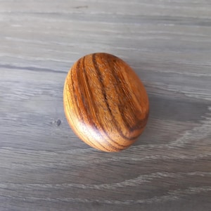 Peut inclure: Objet en bois ovale et lisse, avec une finition polie. Le veinage du bois présente des bandes alternées de brun clair et foncé, créant un motif rayé. Il s'agit probablement d'un objet décoratif ou tactile.