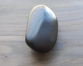 May include: A smooth, dark gray stone with a polished surface. The stone has an oval shape and reflects light, creating highlights. The stone is set against a light gray wooden surface.