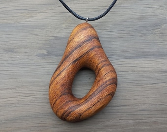 May include: A wooden pendant with a teardrop shape and a large central opening. The pendant is made of brown wood with dark brown grain patterns. It hangs from a black cord.