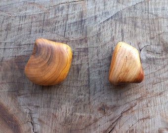 May include: Two smooth, wooden, heart-shaped stones with a natural wood grain pattern. The stones are a light brown color and are sitting on a wooden surface.