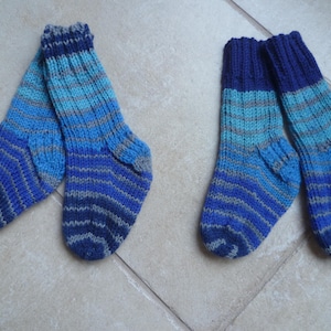 May include: Two pairs of knitted socks in shades of blue and grey. The socks have a striped pattern, with darker blue at the toes and cuffs, transitioning to lighter blues and grey stripes. The socks are handmade.