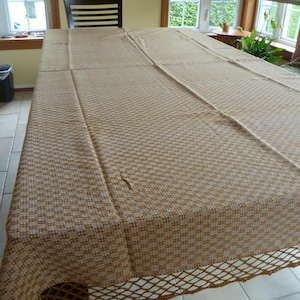 Peut inclure: Une nappe rectangulaire tissée en beige et crème, avec un motif texturé. La nappe a une bordure ajourée décorative. La table est dressée dans une pièce lumineuse avec des fenêtres.