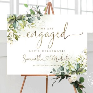 May include: White and green floral engagement announcement sign with gold lettering. The sign reads "We are engaged" and "Let's celebrate! Samantha & Michael" with the date "Saturday, August 12, 2020".
