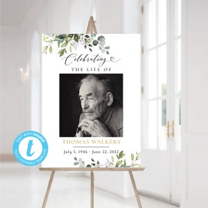以下が含まれることがあります： 男性の白黒写真が載った白い看板。看板には緑と白の花のイラストと「Celebrating the Life of Thomas Walkers July 5, 1946 - June 22, 2022」というテキストが書かれている。