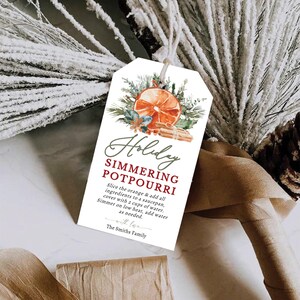 以下が含まれることがあります： オレンジのスライス、シナモンスティック、緑色の水彩画が描かれた白いギフトタグ。タグには「Holiday Simmering Potpourri」というテキストと、シマーリングポプリの作り方の説明が書かれています。タグは麻ひもで結ばれています。