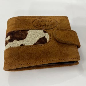 May include: A brown suede bi-fold wallet featuring a central horizontal panel of brown and white cowhide hair-on leather. The front flap includes a snap closure and an embossed oval stamp with the text Couro Legítimo.