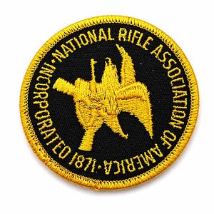 May include: Embroidered patch featuring the words "NATIONAL RIFLE ASSOCIATION OF AMERICA INCORPORATED 1871" in gold lettering around a black circle. The center design is a gold emblem of a rifle and eagle.