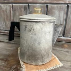 May include: Vintage aluminum coffee pot with a black handle and a clear, amber-colored knob on the lid. The pot sits on a beige, square coaster with a lace trim. The background features weathered wooden panels.
