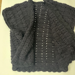 May include: A dark charcoal-colored, open-front crochet cardigan. The handmade garment features a scalloped edge and a textured stitch pattern. The cardigan has short sleeves and a relaxed fit, suitable for layering. The open design allows for versatile styling.