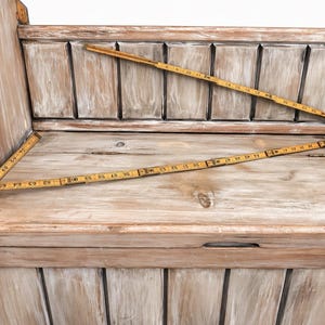 May include: A light-colored wooden bench with a distressed finish. Three vintage folding rulers are laid across the bench, with measurements in inches. The bench has a slatted back and a storage compartment.