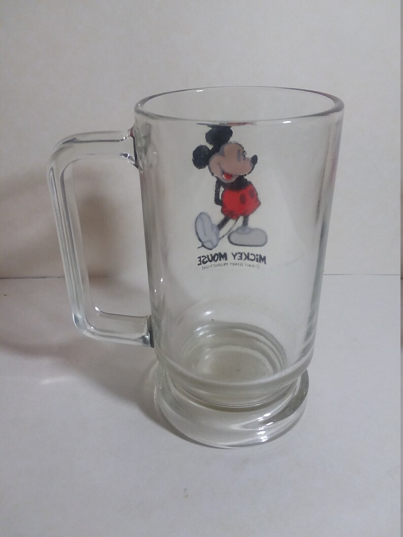 May include: A clear glass mug with a Mickey Mouse design. The mug has a handle and a wide base. The text "Walt Disney Productions" and "Mickey Mouse" are printed on the mug.