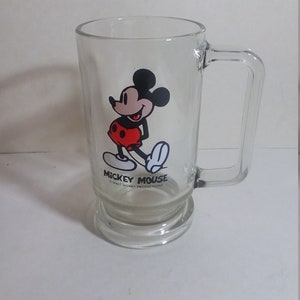 May include: A clear glass mug with a Mickey Mouse design. The mug has a handle and a wide base. The design features Mickey Mouse in his classic red shorts and yellow shoes. The text "Mickey Mouse" and "Walt Disney Productions" is printed on the mug.