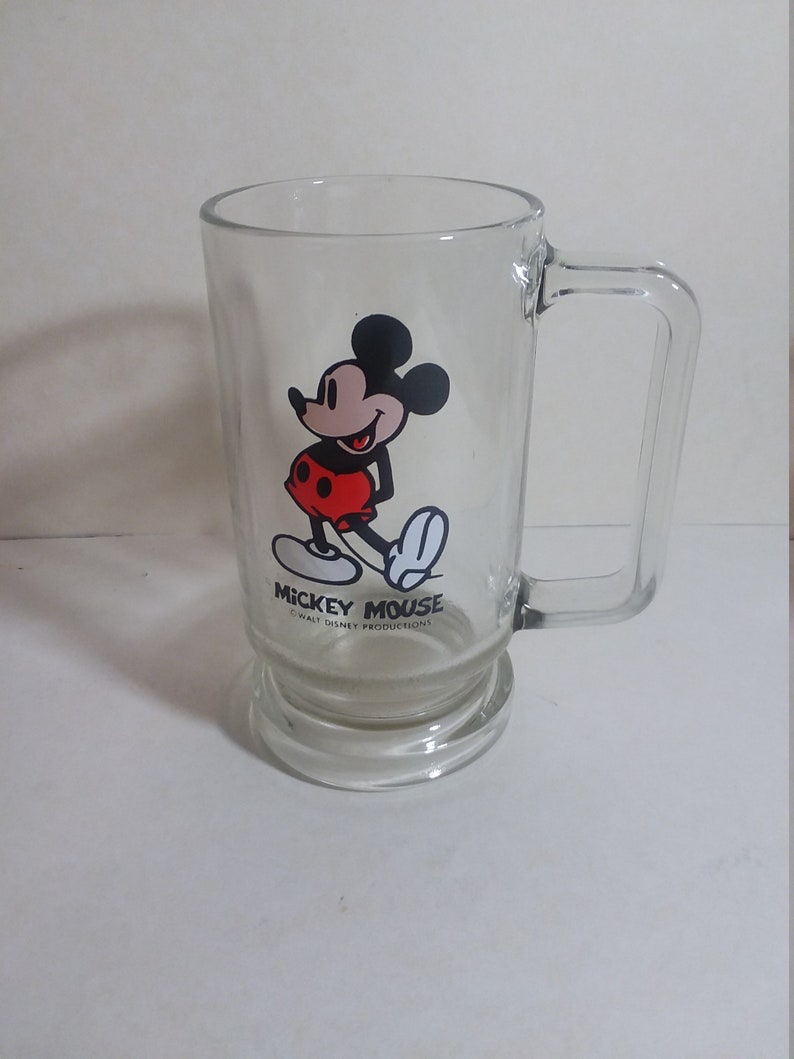May include: A clear glass mug with a Mickey Mouse design. The mug has a handle and a wide base. The design features Mickey Mouse in his classic red shorts and yellow shoes. The text "Mickey Mouse" and "Walt Disney Productions" is printed on the mug.