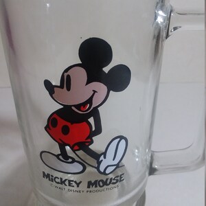 May include: A clear glass mug with a black and white cartoon image of Mickey Mouse wearing red shorts. The mug has a handle and the text "Mickey Mouse" and "&copy; WALT DISNEY PRODUCTIONS" printed on the bottom.
