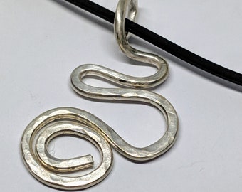 Puede incluir: Un collar con un colgante de plata con un diseño abstracto martillado. El colgante tiene forma de una serie de bucles y curvas, y cuelga de un cordón negro.