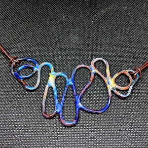 May include: A blue, red, and yellow abstract metal necklace with a brown leather cord.