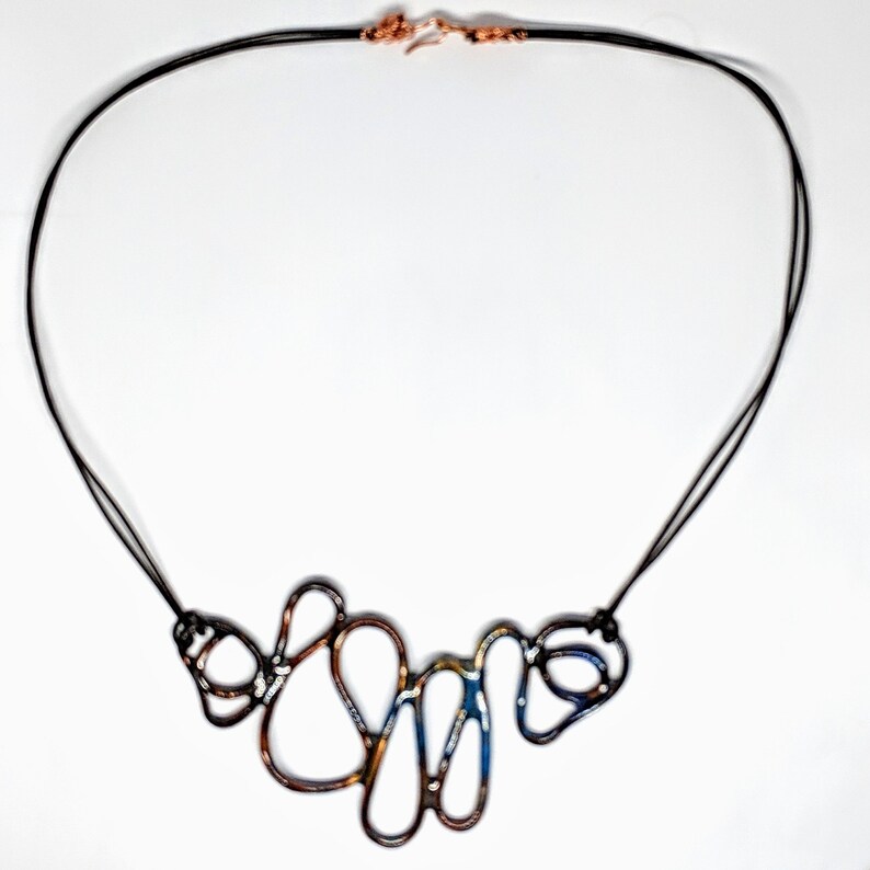May include: A necklace with a long black cord and a multi-colored metal pendant. The pendant is made of several interconnected loops of metal, with a blue, gold, and copper finish.