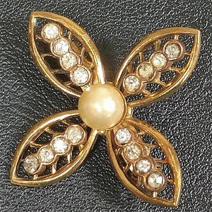 May include: A gold-tone brooch in the shape of a four-leaf clover with a faux pearl in the center. Each leaf is decorated with clear rhinestones.