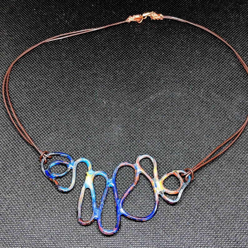 May include: A blue, yellow, and brown abstract metal necklace with a brown leather cord.