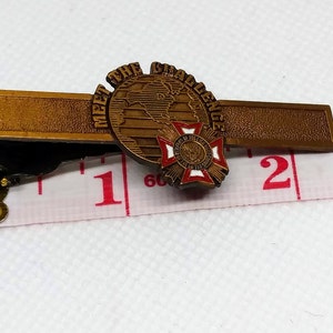 Op de afbeelding: Een gouden stropdasclip met een wereldbol en een rood kruis. De wereldbol heeft de tekst "Meet the Challenge" eromheen. De stropdasclip is ongeveer 2,5 cm lang.