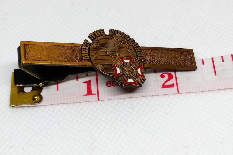 Op de afbeelding: Een gouden stropdasclip met een wereldbol en een rood kruis. De wereldbol heeft de tekst "Meet the Challenge" eromheen. De stropdasclip is ongeveer 2,5 cm lang.