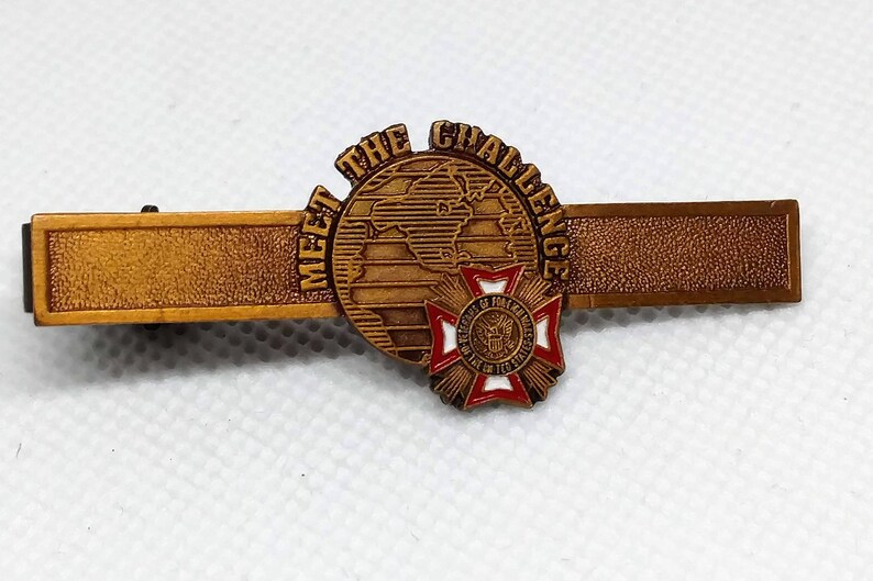Op de afbeelding: Gouden stropdasgesp met een wereldbol en de woorden "Meet the Challenge". De wereldbol is omgeven door een rood-wit-blauw kruis met een zegel in het midden.