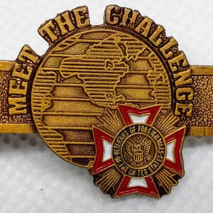 Op de afbeelding: Vergulde revers-speld met de woorden "Meet the Challenge" rond een wereldbol. De speld heeft ook een rood-wit-blauw kruis met de woorden "Veterans of Foreign Wars of the United States" in een cirkel.