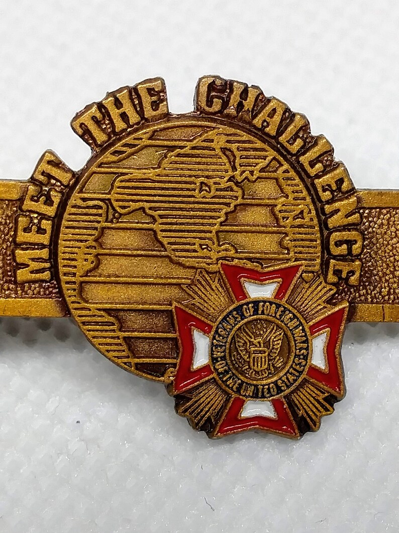 Op de afbeelding: Vergulde revers-speld met de woorden "Meet the Challenge" rond een wereldbol. De speld heeft ook een rood-wit-blauw kruis met de woorden "Veterans of Foreign Wars of the United States" in een cirkel.