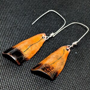 May include: A pair of copper earrings with a black and orange design. The earrings are shaped like a teardrop and have a silver hook.