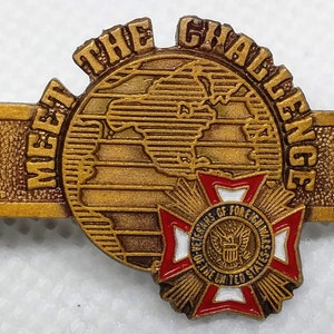 Op de afbeelding: Gouden metalen pin met de woorden "Meet the Challenge" rond een wereldbolontwerp. De wereldbol is gedeeltelijk bedekt door een rode, witte en blauwe stervormige pin met de tekst "Veterans of Foreign Wars of the United States".