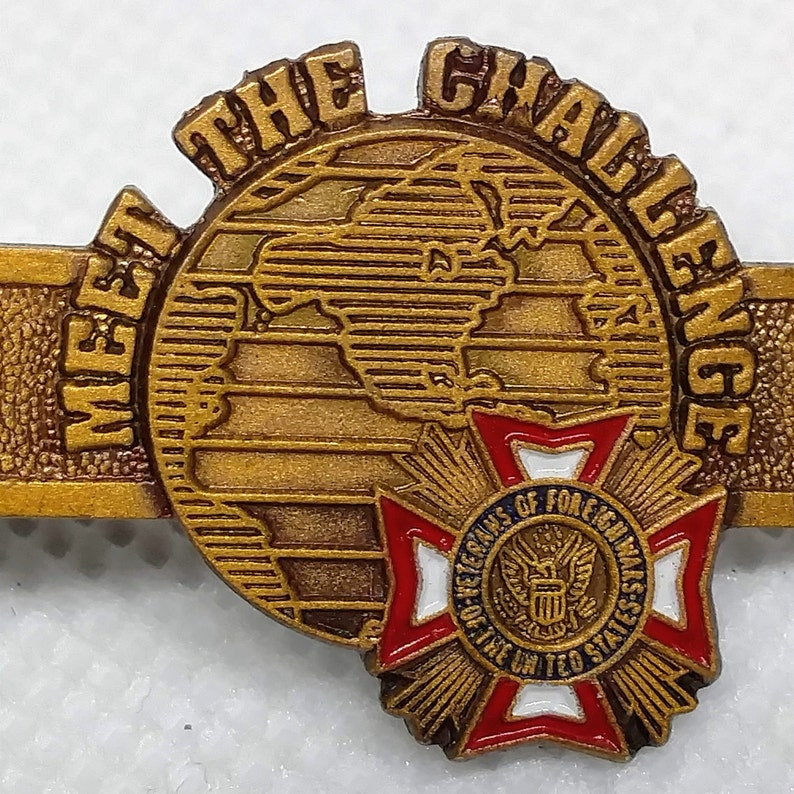 Op de afbeelding: Gouden metalen pin met de woorden "Meet the Challenge" rond een wereldbolontwerp. De wereldbol is gedeeltelijk bedekt door een rode, witte en blauwe stervormige pin met de tekst "Veterans of Foreign Wars of the United States".