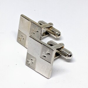 May include: A pair of silver cufflinks with a geometric design. Each cufflink is square shaped with a pattern of lines and a small design in the centre.