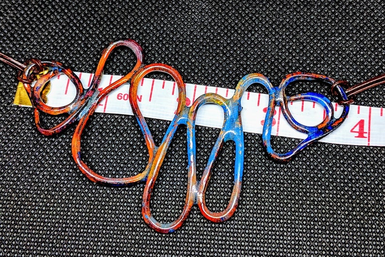 May include: A colorful copper wire necklace with blue, orange, and yellow enamel paint. The necklace is approximately 3 inches long and is measured with a tape measure.