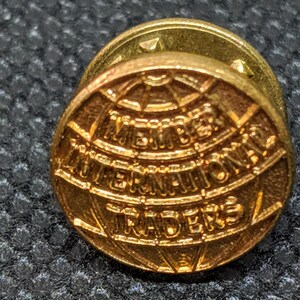 Könnte beinhalten: Eine goldfarbene Anstecknadel mit einem Globus-Design und dem Text "Member International Traders".