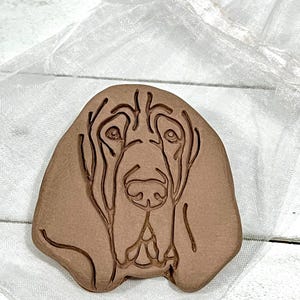 May include: A brown, dog-shaped ceramic ornament featuring a detailed relief of a Bloodhound's face. The ornament is presented on a white, sheer fabric, suggesting it may be a gift or decorative item. The dog's features are clearly defined.