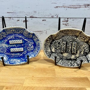 May include: Two ceramic dishes with intricate designs. One dish is blue and white with a floral pattern and the other is black and white with a paisley pattern. Both dishes have a raised design of three lines.
