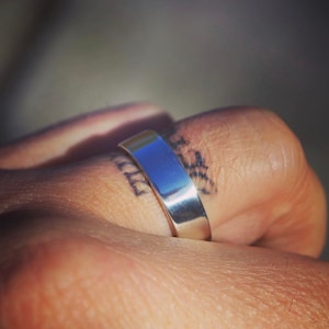 May include: A silver band ring worn on a finger with a tattoo.