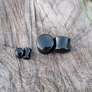 May include: Three black ear gauges of different sizes. The largest gauge is a cylinder shape, while the two smaller gauges are round.