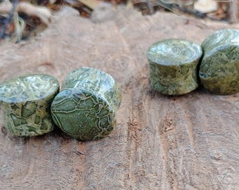 May include: Six green stone plugs with a textured, patterned surface. The plugs are cylindrical with a rounded top and bottom. They are arranged on a wooden surface.
