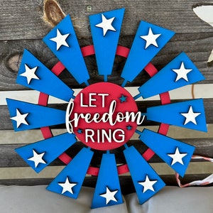 May include: A red, white, and blue wooden wind spinner with the text "Let freedom ring" in the center. The wind spinner has 12 blades with white stars on each blade.