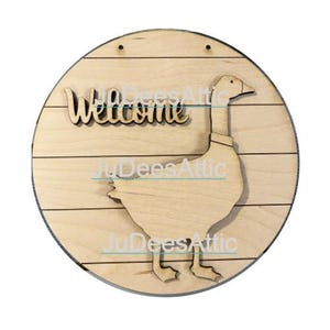 Könnte beinhalten: Ein rundes Holzschild mit rustikalem Holzmaserungsmuster. Das Schild zeigt eine ausgeschnittene Gans mit dem Wort "Willkommen" darüber.