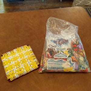 May include: A yellow and white woven trivet with a dark brown border is next to a clear bag filled with colourful loops. The bag contains loops in various colours, possibly for crafting or weaving projects.