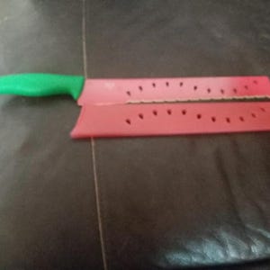 May include: A red and green watermelon-themed knife with a matching sheath. The knife has a green handle and a red blade with black dot details. The sheath is red with black dot details.
