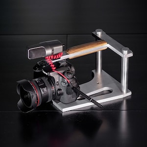 May include: A black camera with a wide-angle lens is mounted on a silver metal tripod with a wooden handle. A black microphone is attached to the top of the camera.
