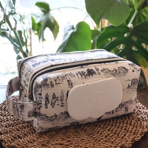 May include: A rectangular fabric pouch featuring a black and white toile print of desert landscapes with cacti and figures on horseback. The pouch includes a white plastic flip-top dispenser lid on the front panel and a side carry handle.