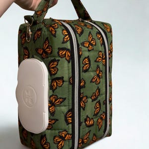 Könnte beinhalten: Gesteppte grüne Tasche mit einem Muster aus orangefarbenen und schwarzen Schmetterlingen. Die Tasche hat ein weißes, rechteckiges Fach und einen Griff. Sie verfügt über silberne Reißverschlüsse und einen abnehmbaren Riemen.